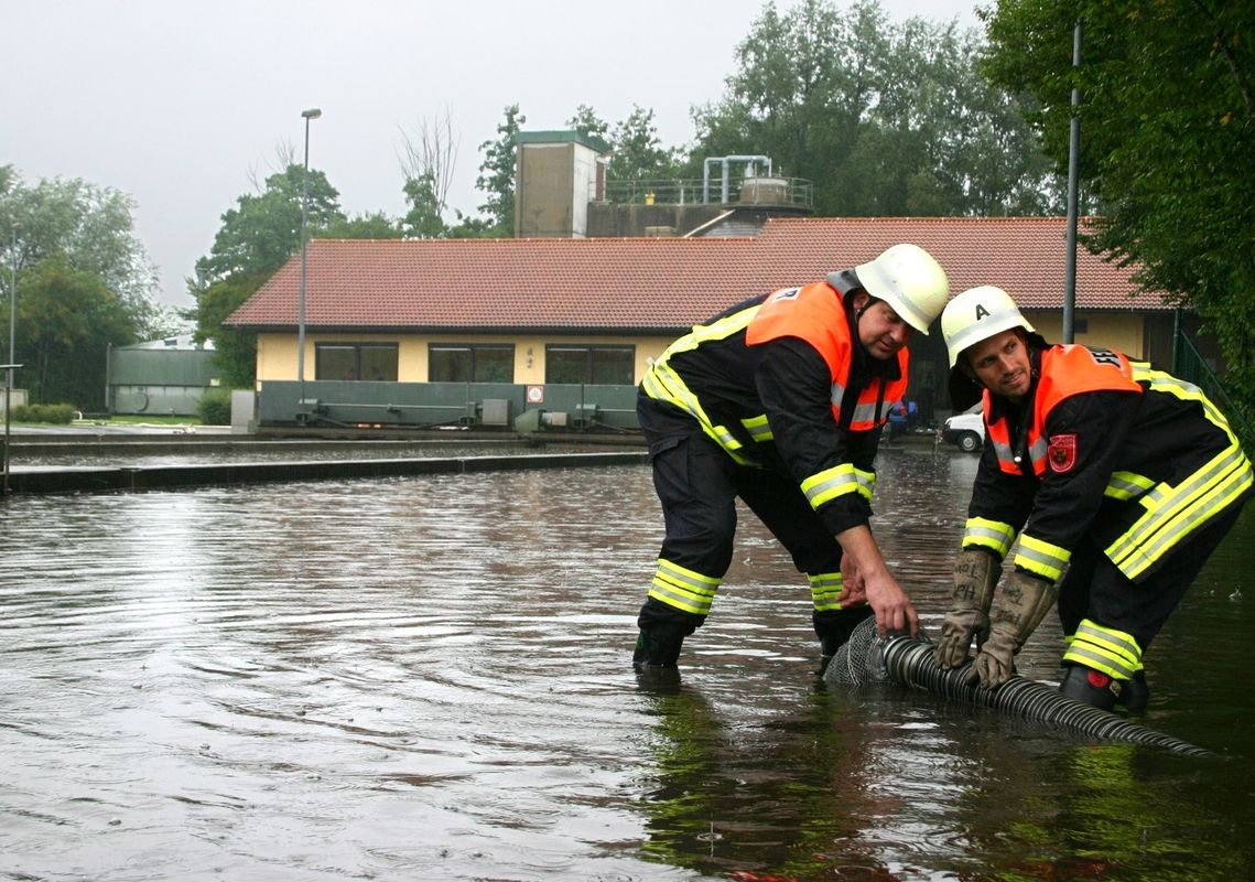 Zwei Feuerwehrleute in Schutzkleidung und Helmen stehen knietief im Wasser und heben gemeinsam einen Gegenstand