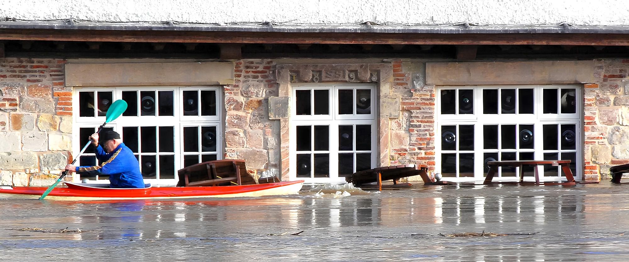 Person in blauem Oberteil sitzt in einem roten Kajak auf ruhigem Wasser vor einem Gebäude mit drei weißen Fenstern und Steinmauer