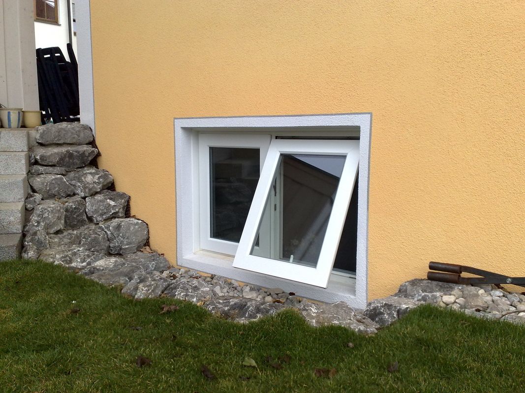 Kellerschachtfenster mit einem geöffneten weißen Rahmen in einer gelben Hauswand, umgeben von Steinen und Rasen